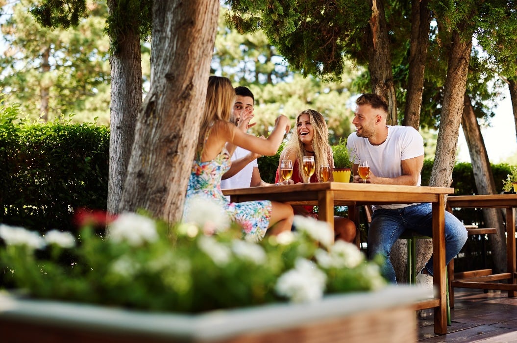 Eine Gruppe junger Leute sitzt an einem Tisch in einer Sommerbar, lacht und trinkt Bier